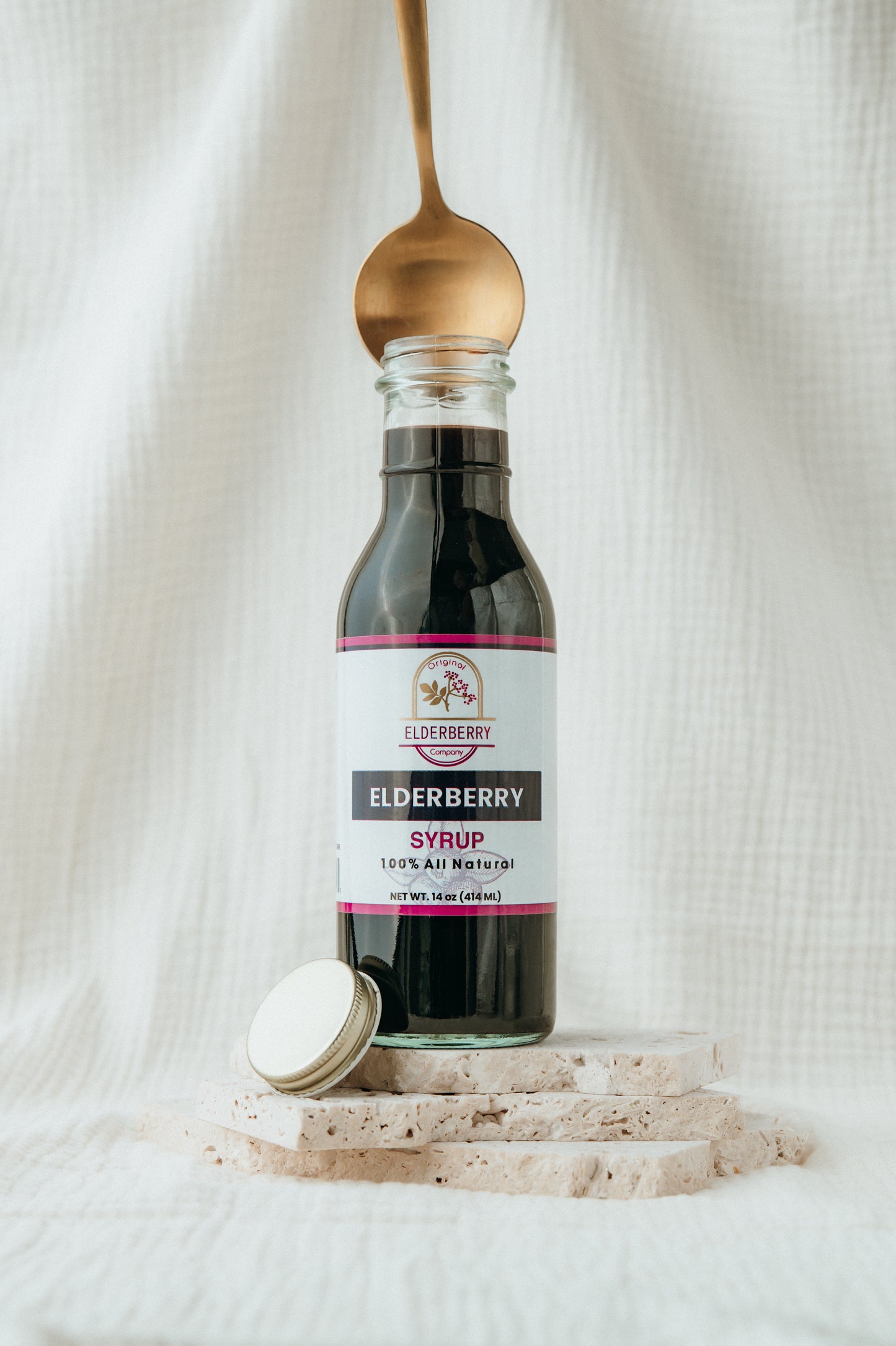 Bottle of elderberry syrup with a gold spoon on a white fabric background