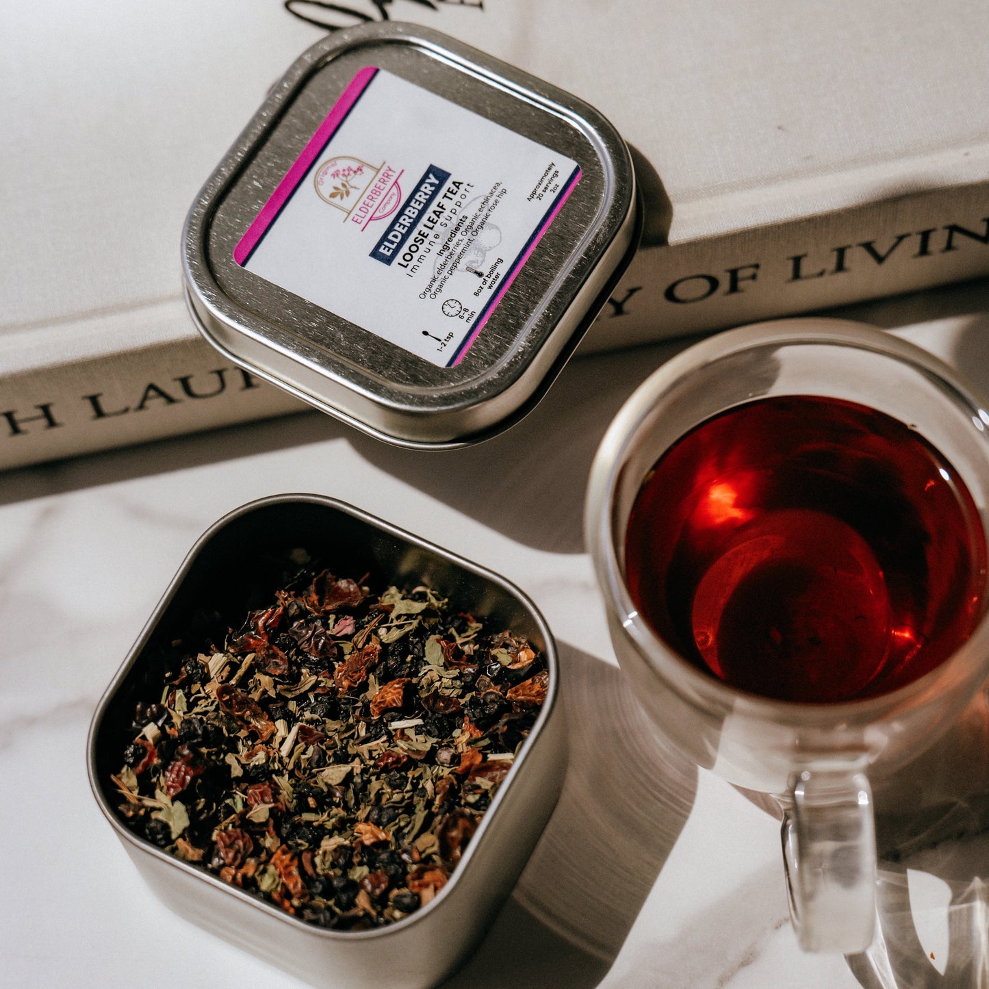 Tea tin with tea leaves, a cup of tea, and a book on a marble surface.