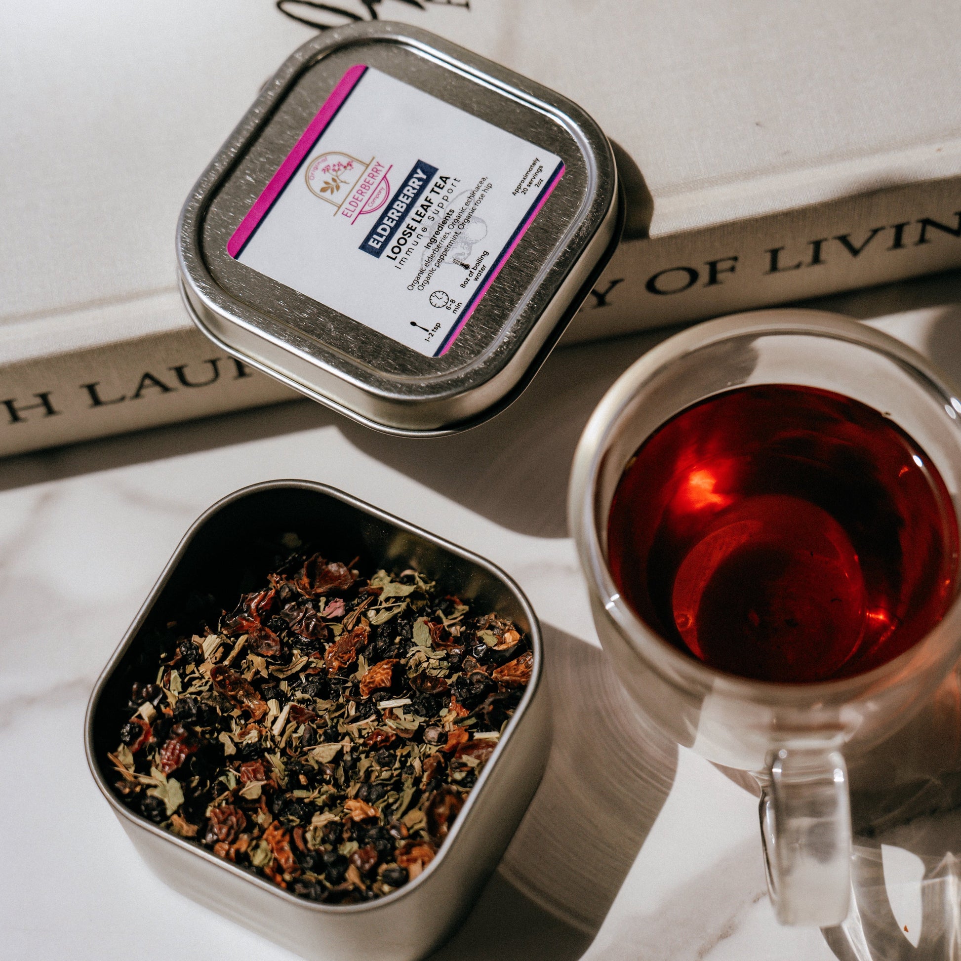 Tea tin with tea leaves, a cup of tea, and a book on a marble surface.
